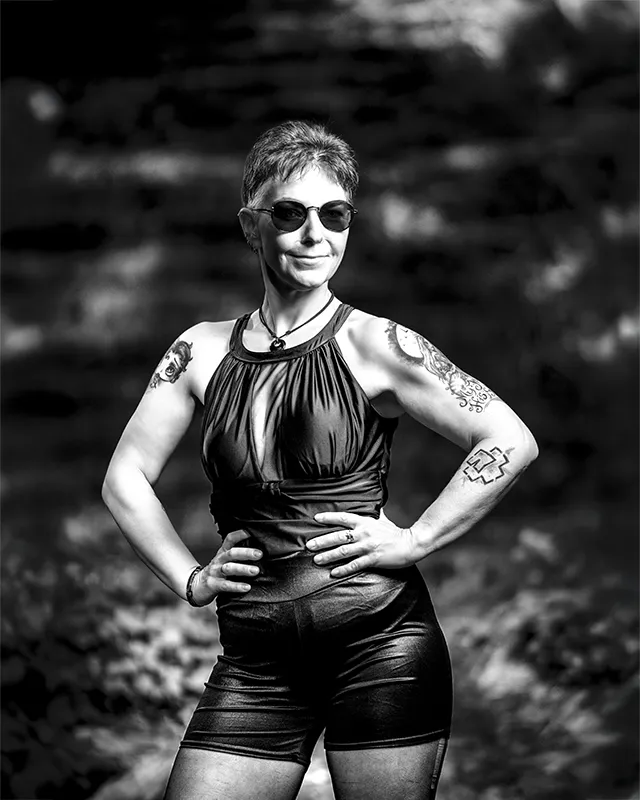 Schwarz-Weiß-Porträt einer tätowierten Frau mit Sonnenbrille und kurzem Haar in natürlicher Umgebung.