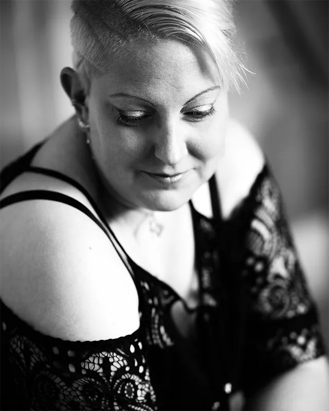 Schwarz-Weiß-Porträt einer Frau mit kurzem Haar und Spitzenoberteil, die mit gesenktem Blick lächelt.